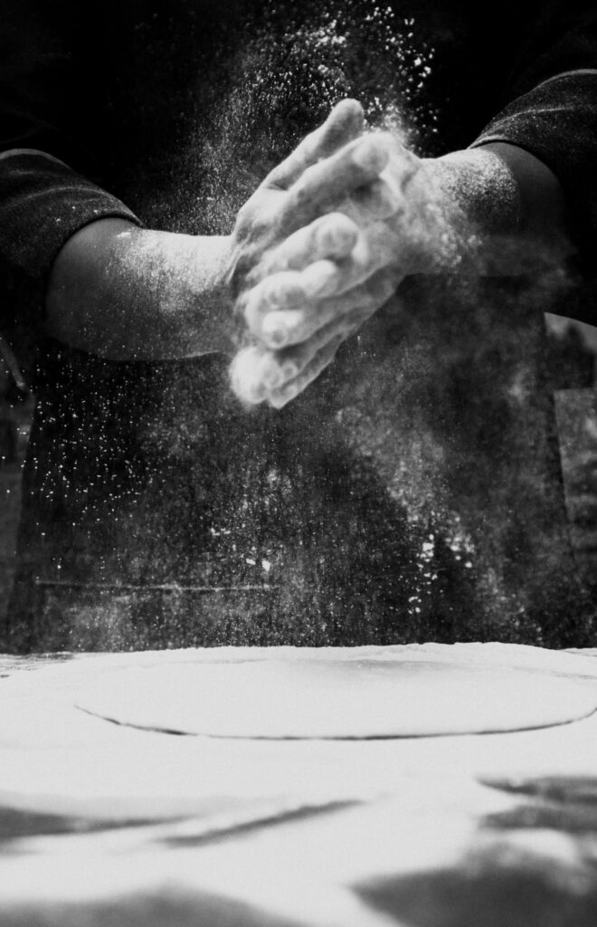 Black and white photo of a chef clapping flour in a kitchen, creating a dramatic effect.
